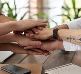 7 Hände, die übereinandergelegt sich als Zeichen des Zusammenhalts über eienr Tischplatte, auf der Smartphones und Notizbücher liegen.